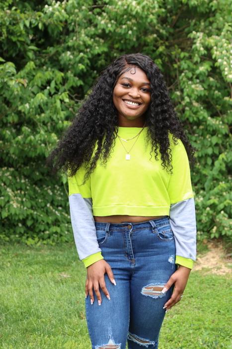 Ama, smiling and wearing a bright green shirt and blue jeans with trees behind her. 